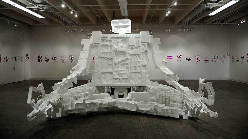 An art gallery, with pictograms, emojis, and symbols running in a line around the walls of the room. In the center of the room there is a massive robotic figure made of styrofoam, intensely detailed with joints, panels, pistons, and wires. The "Styrobot" is in a classic "lotus" meditation pose: legs crossed and arms on the knees.