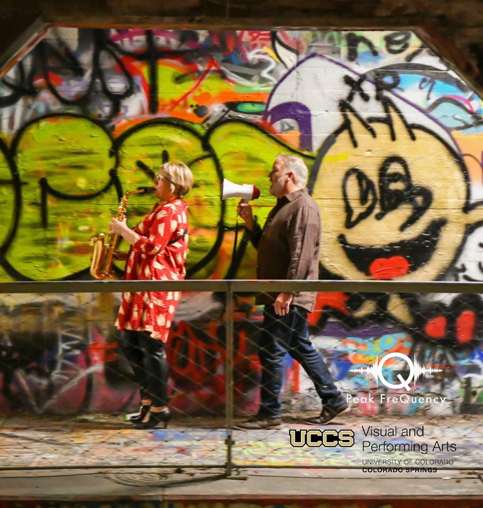 Bent Frequency Duo's Jan Berry Baker with a saxophone and Stuart Gerber with a megaphone walking in front of a wall painted with street art